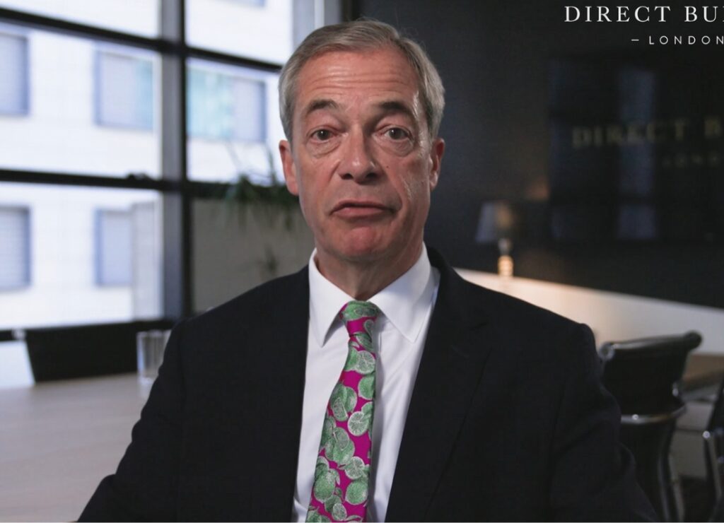 Nigel Farage speaking during an indoor interview, wearing a dark suit and colourful patterned tie.
