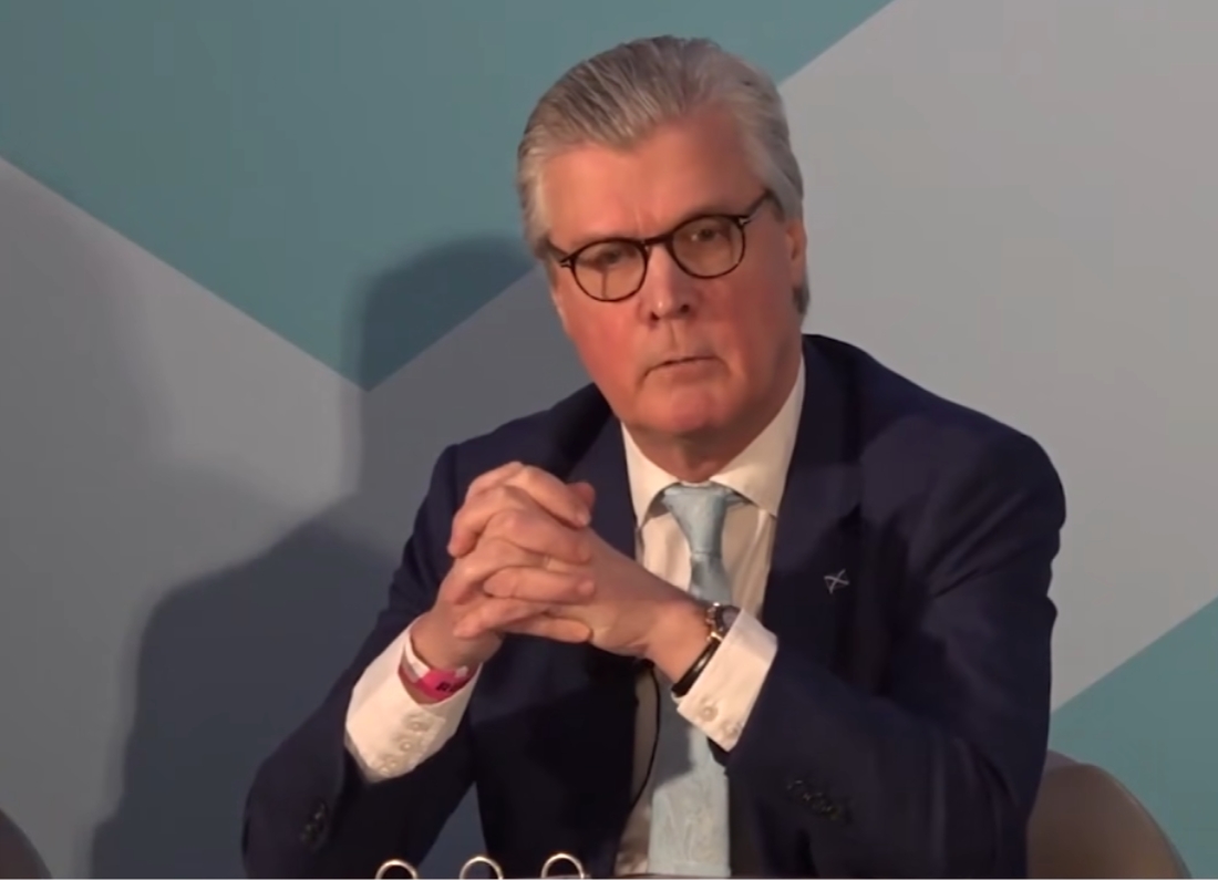 Malcolm Offord sits on a panel with hands clasped, speaking during a formal event.