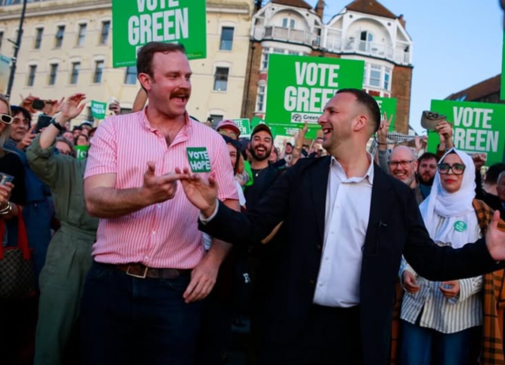 Rob Yates and Green Party leader Zack Polanski laughing and celebrating with supporters holding "Vote Green" signs during a 2026 local election campaign event.