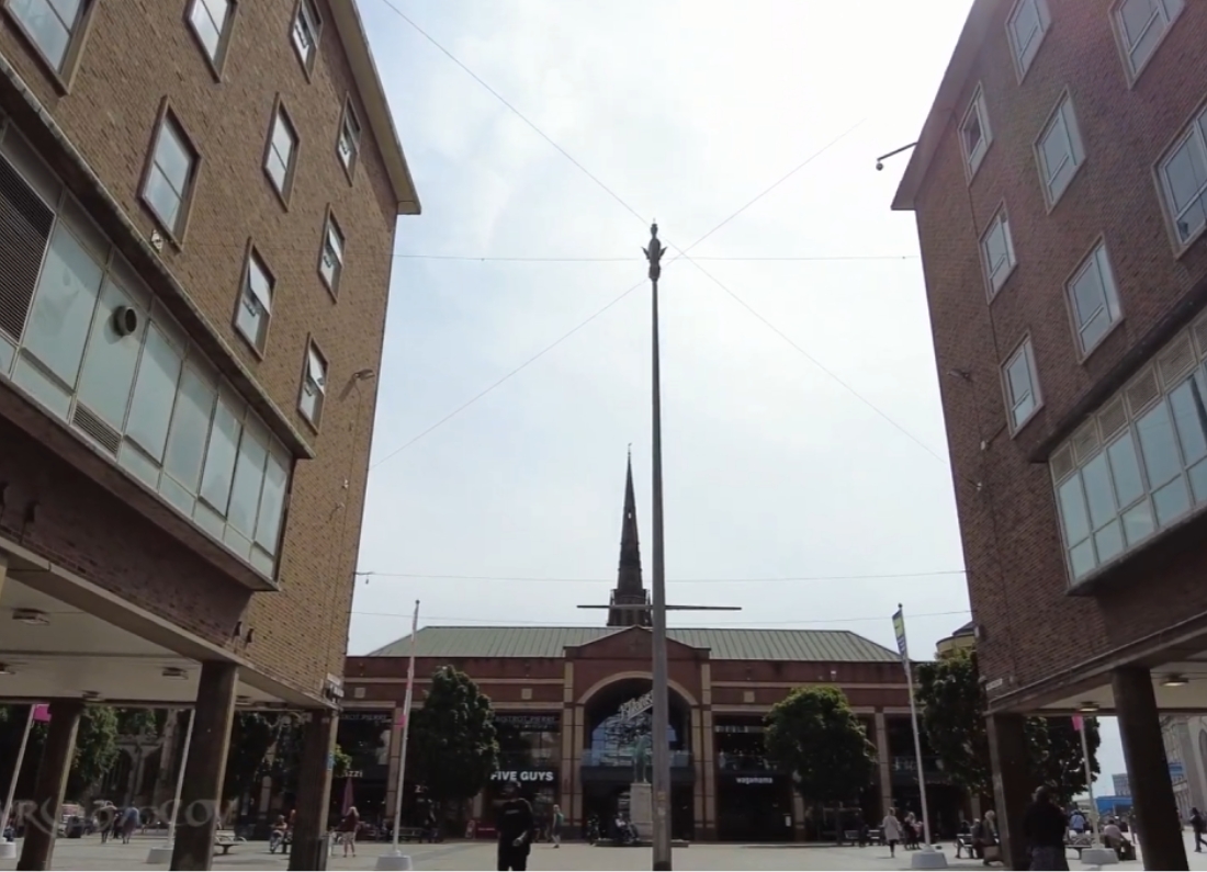 View through Coventry city centre shopping area toward a church spire, framed by two brick buildings.