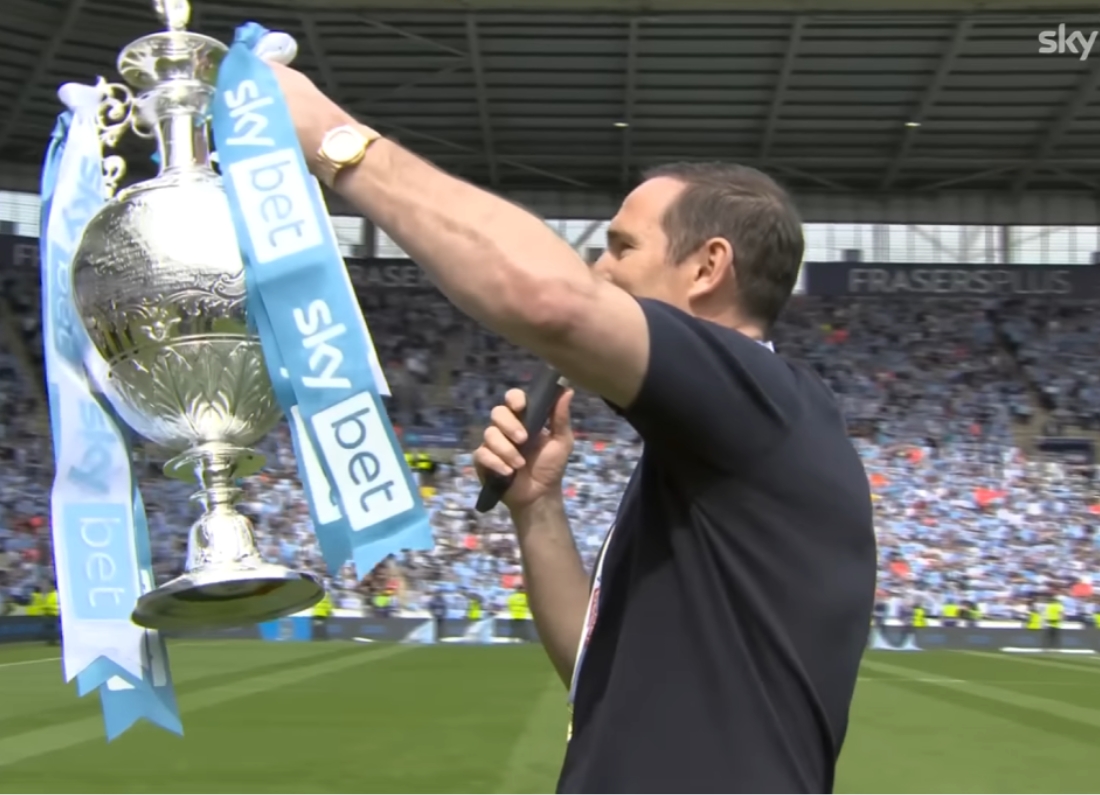 Frank Lampard holds the Championship trophy while speaking to Coventry City fans on the pitch.