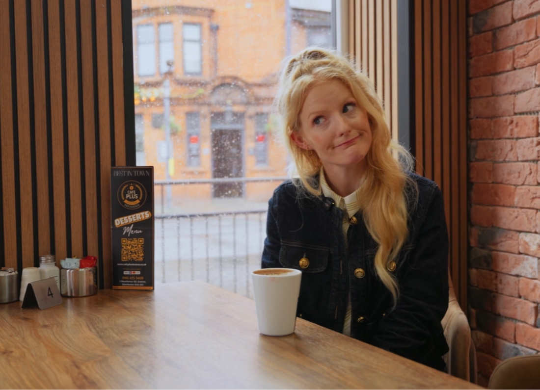 Hannah Spencer sits in a café by the window with a cup of coffee, smiling while looking to the side.