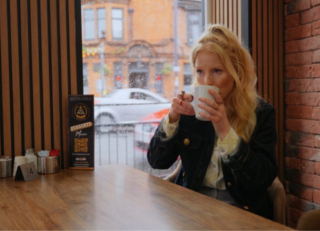 Hannah Spencer sits in a café by the window drinking from a white mug on a rainy day.