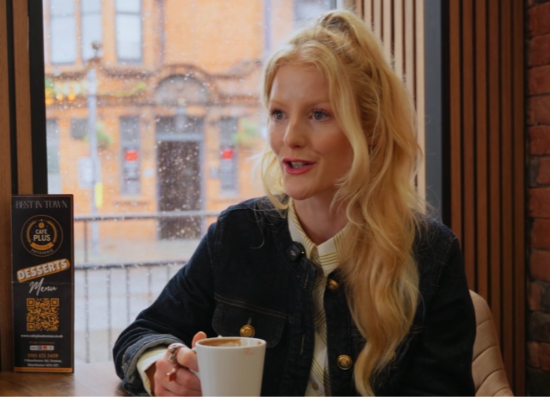 Hannah Spencer sits in a café holding a cup of coffee while speaking, with rain visible on the window behind her.