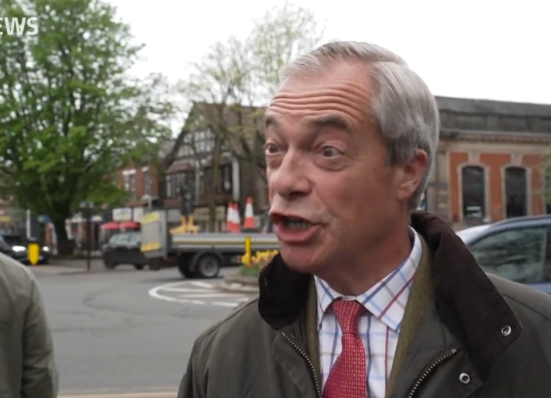 Nigel Farage speaking animatedly outdoors during a local election campaign, wearing a green field jacket, checkered shirt, and red tie.