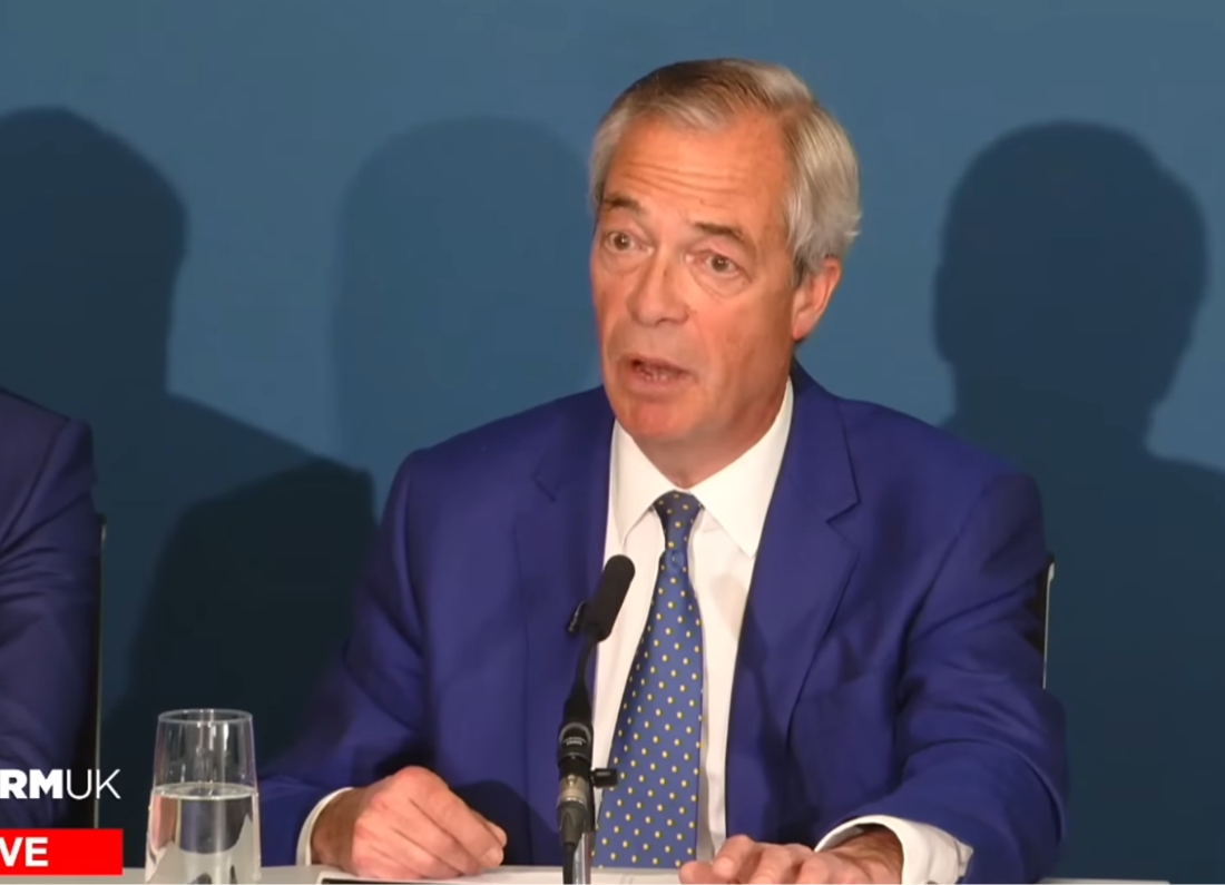 Nigel Farage speaking at a press conference behind a microphone, wearing a blue suit and patterned tie.