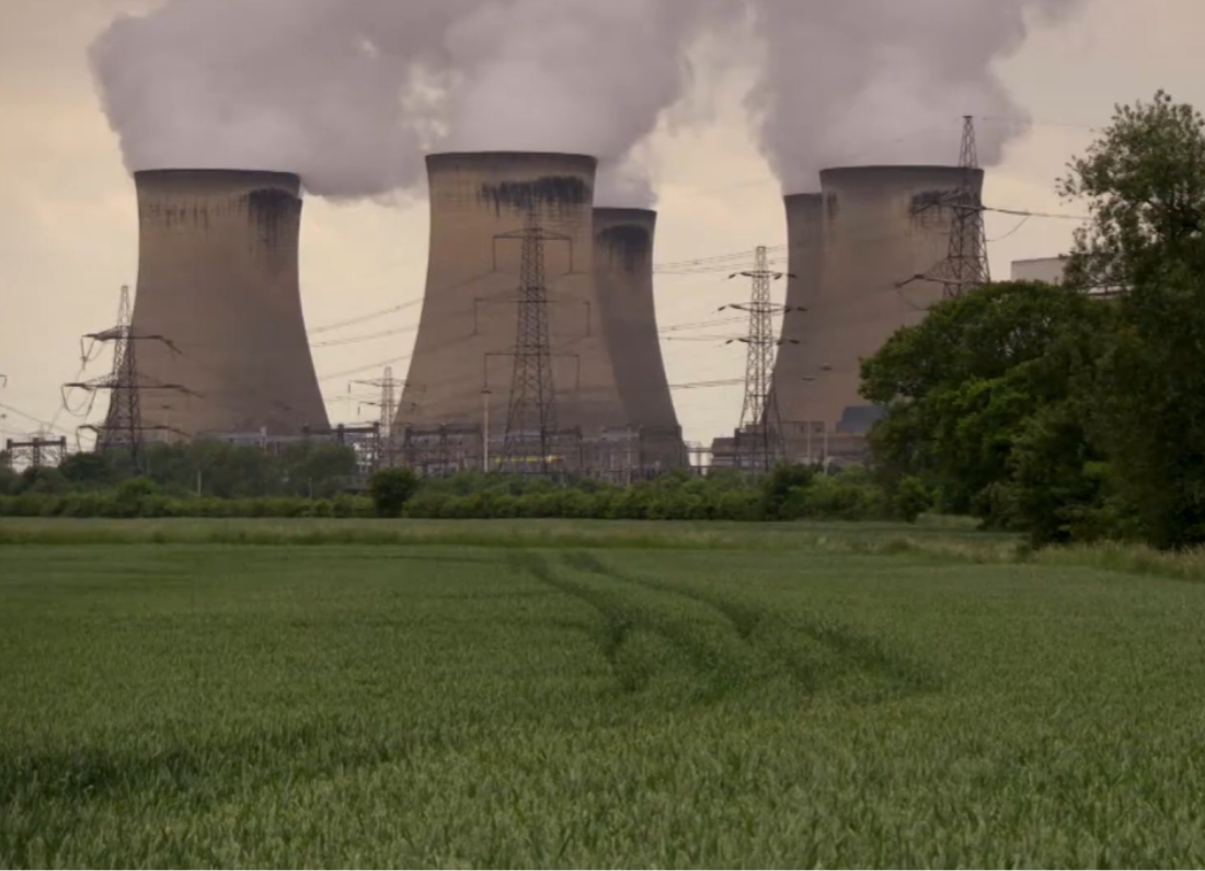 Power station cooling towers emitting steam beside a green field and electricity pylons.