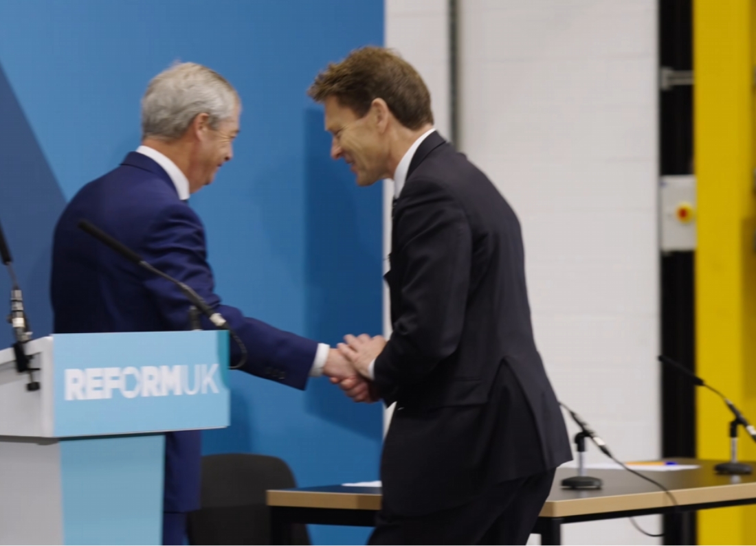 Nigel Farage and Richard Tice shaking hands on stage at a Reform UK event in April 2026, with a Reform UK-branded podium in the foreground against a blue backdrop.