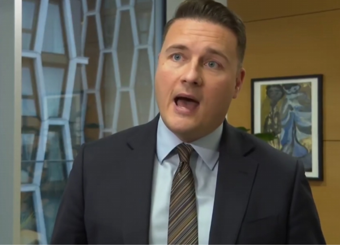 Health Secretary Wes Streeting speaking during an indoor interview in a modern office, wearing a dark suit and striped tie.