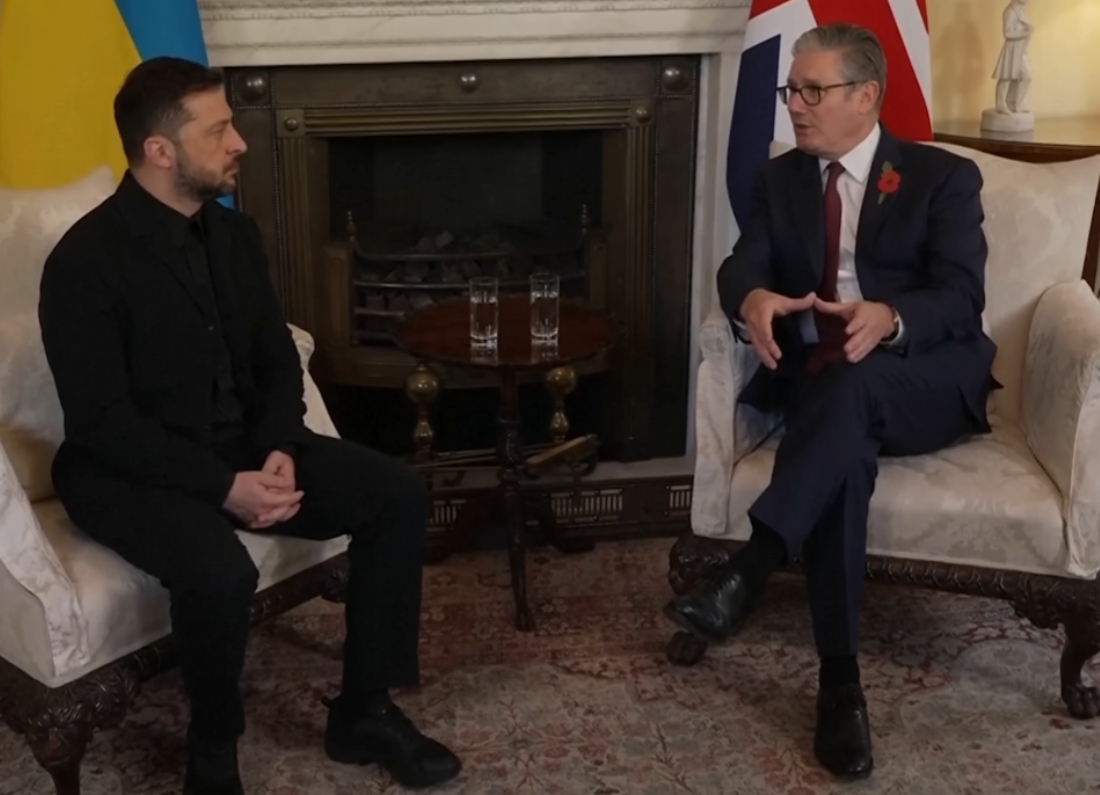 UK Prime Minister Keir Starmer, wearing a dark suit with a red poppy pin, gestures during a discussion with Ukrainian President Volodymyr Zelenskyy, who is dressed in a black tactical shirt, seated in 10 Downing Street.
