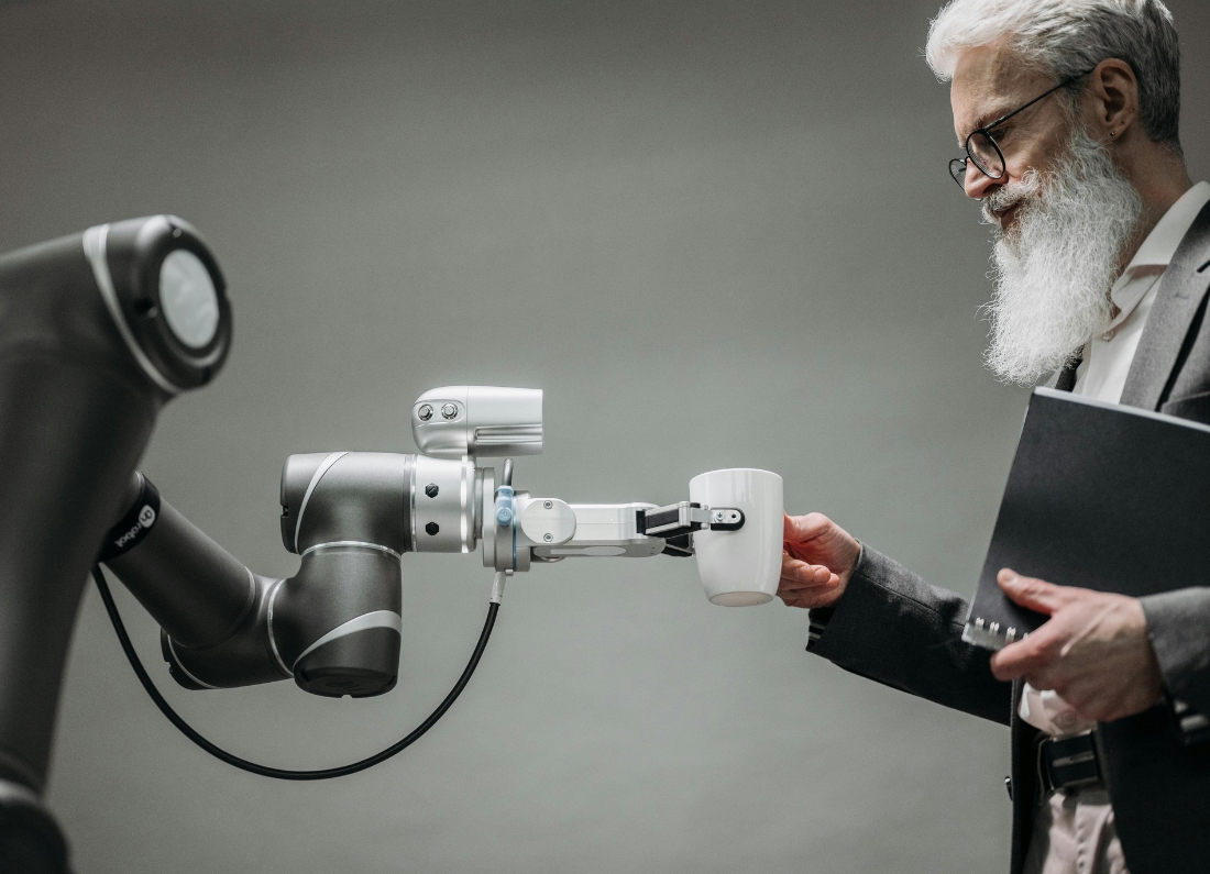 A robotic arm holds out a white coffee mug to a man with a white beard and glasses, who is reaching to take it while holding a notebook. The scene illustrates human interaction with automation and artificial intelligence technology.