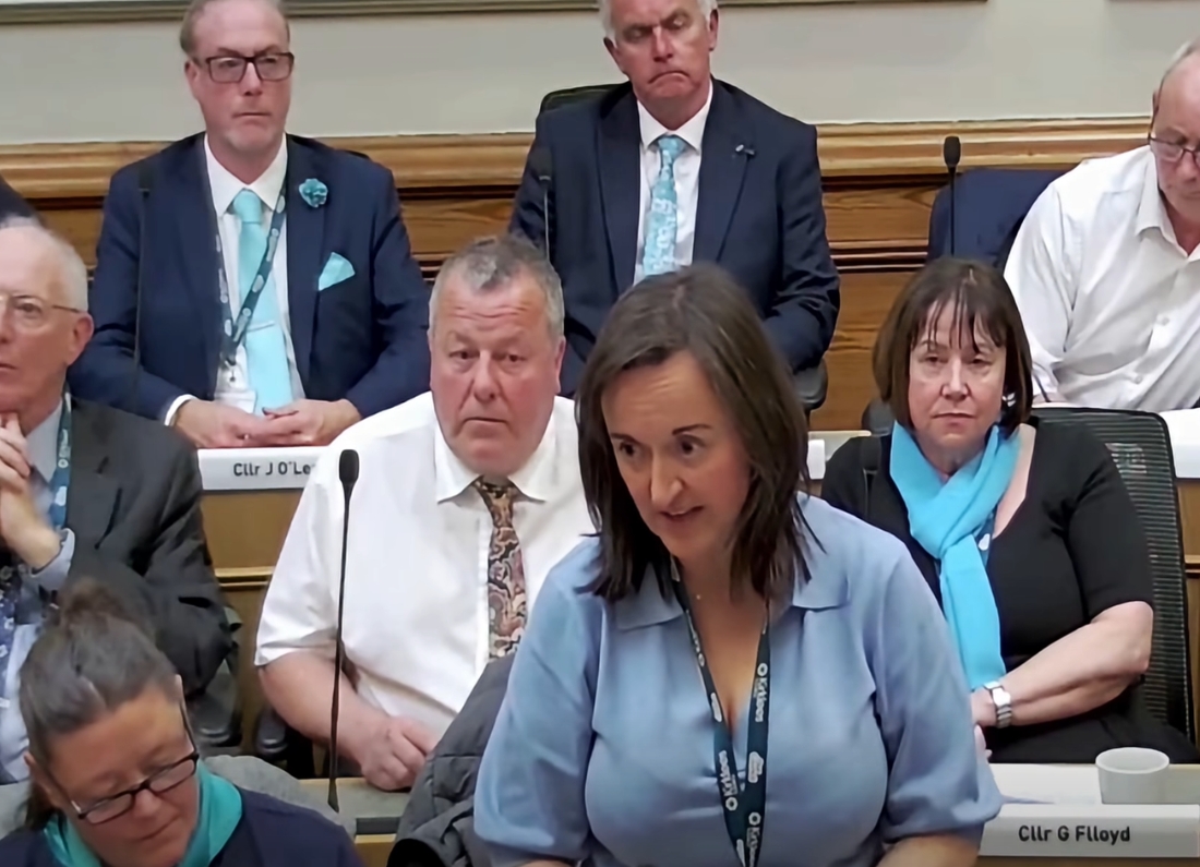 A tense Kirklees Council chamber meeting showing councillors seated during a debate, with Councillor Sarah Wood standing and speaking into the chamber.