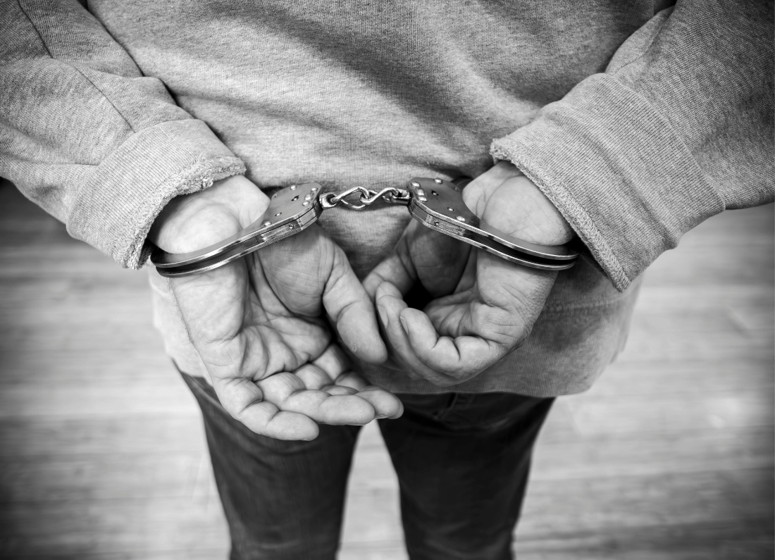 Black-and-white close-up image of a person in a grey hoodie standing with their hands cuffed behind their back.