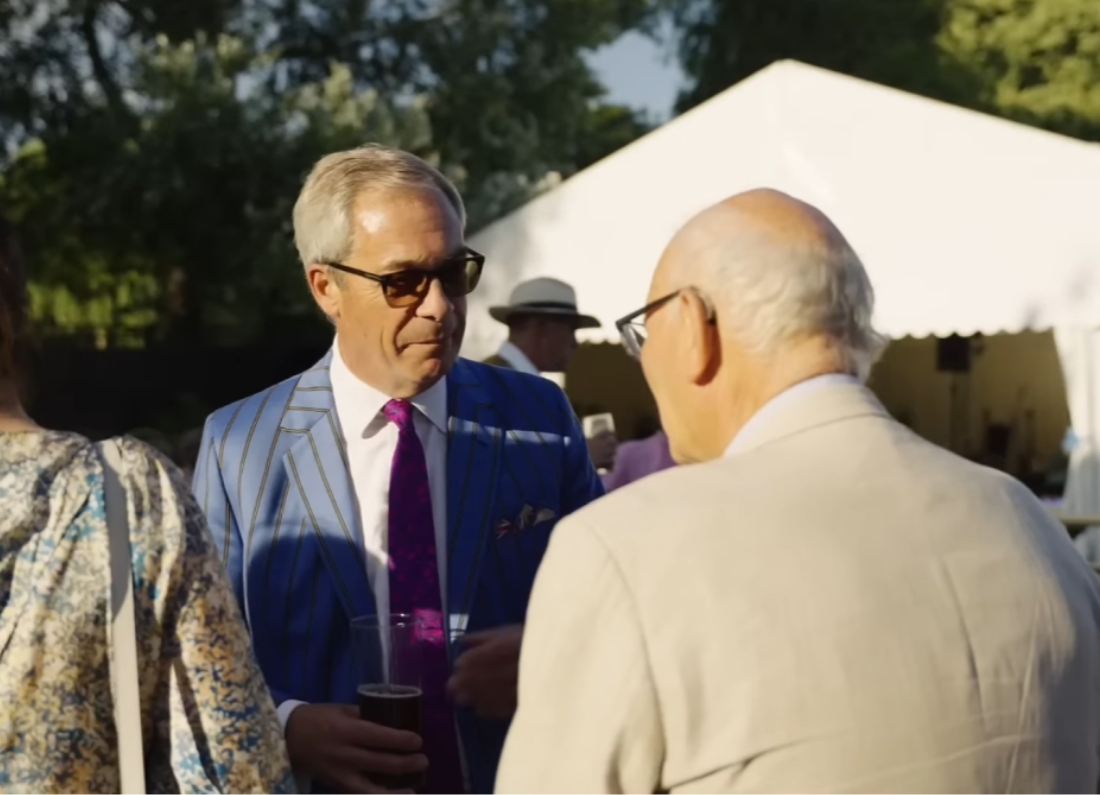 Reform UK leader Nigel Farage speaks with guests at an outdoor event in Clacton while wearing a blue striped suit and sunglasses.