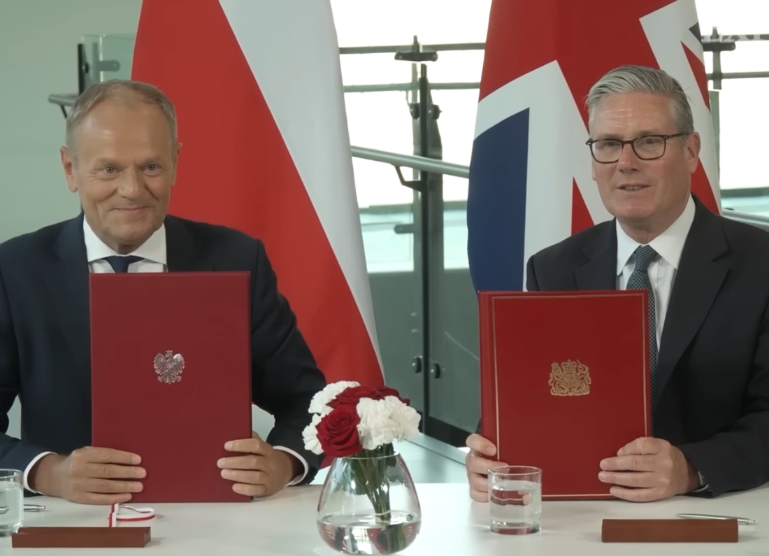 Polish Prime Minister Donald Tusk and UK Prime Minister Keir Starmer sit at a table holding signed red treaty folders during a formal signing ceremony. Polish and British flags stand behind them, with flowers and glasses of water on the table.