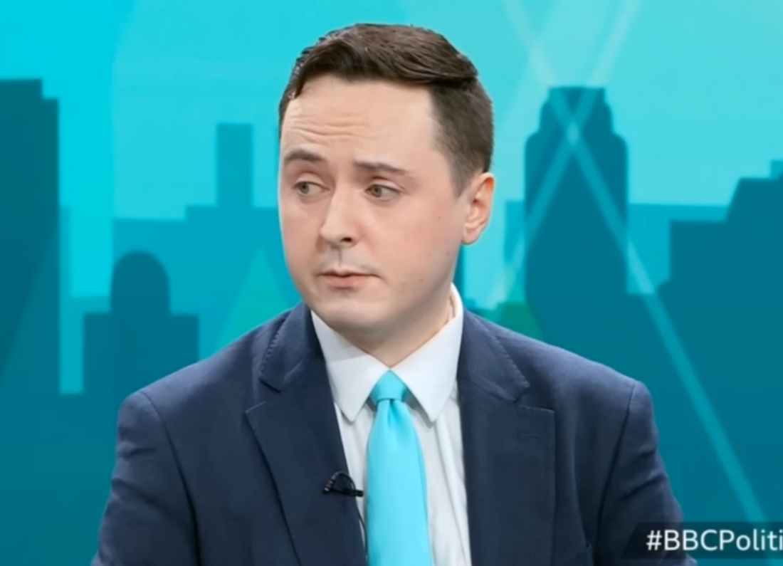 Reform UK councillor Darren Grimes appears during a television interview wearing a navy suit, white shirt and turquoise tie. A stylised city skyline graphic is visible in the blue background.