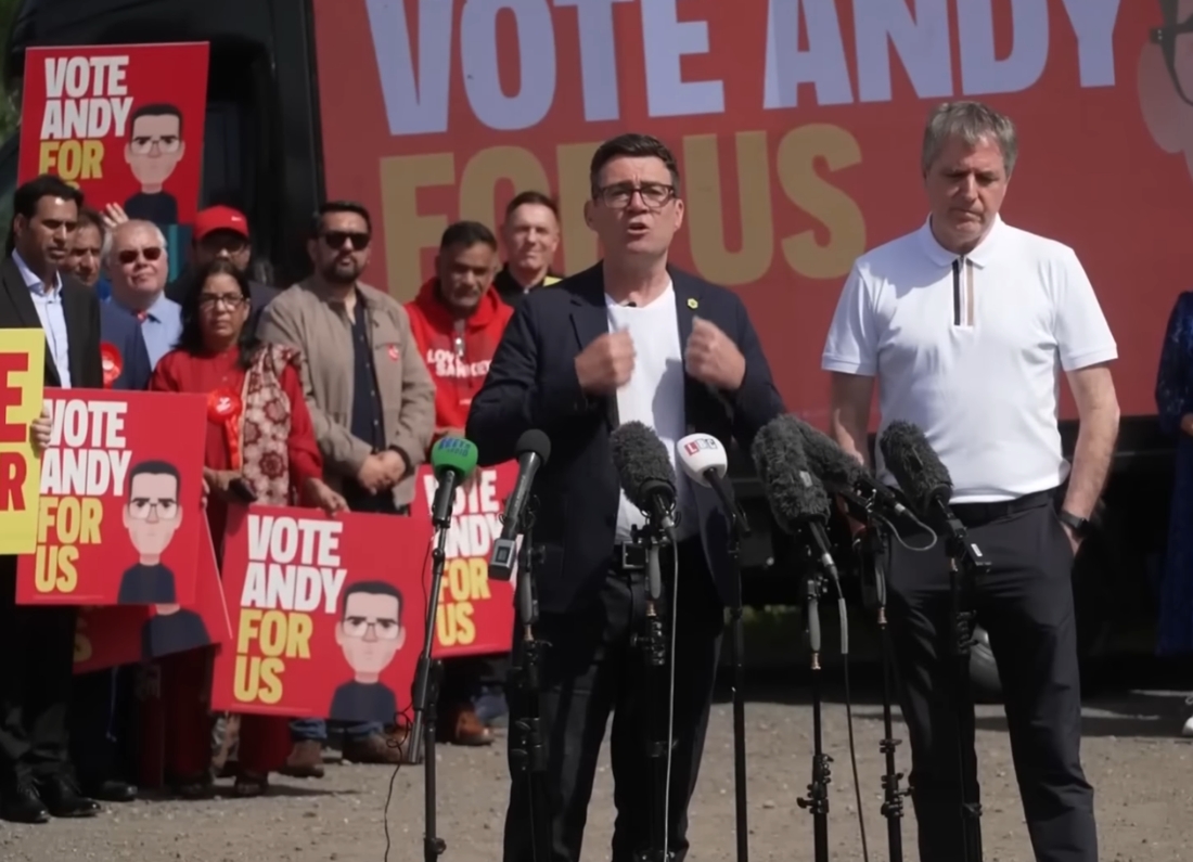 Andy Burnham speaking at an outdoor campaign event surrounded by supporters holding “Vote Andy For Us” signs.
