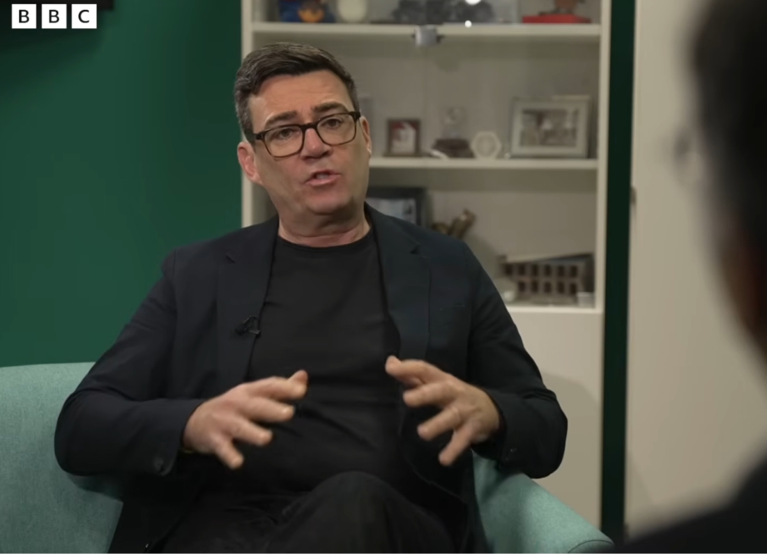 Greater Manchester Mayor Andy Burnham speaking during a BBC interview while seated in a studio chair wearing a black suit and glasses.