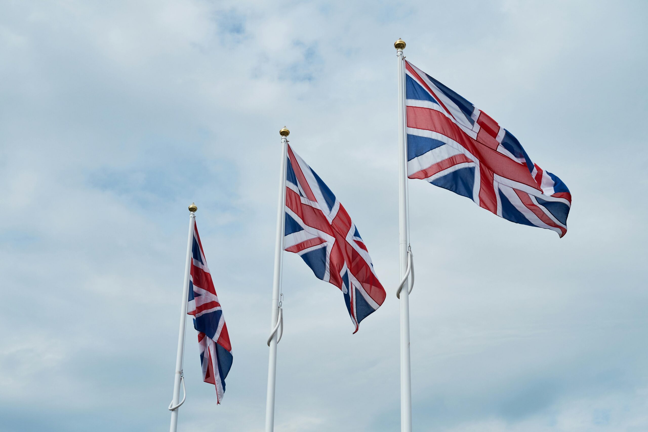 Three Union Jack flags flying on tall white flagpoles against a cloudy sky.