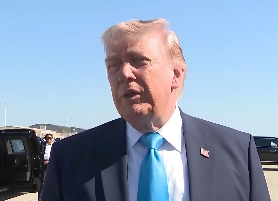 Donald Trump speaking to reporters outdoors near vehicles and airport buildings during a media scrum under a clear blue sky.