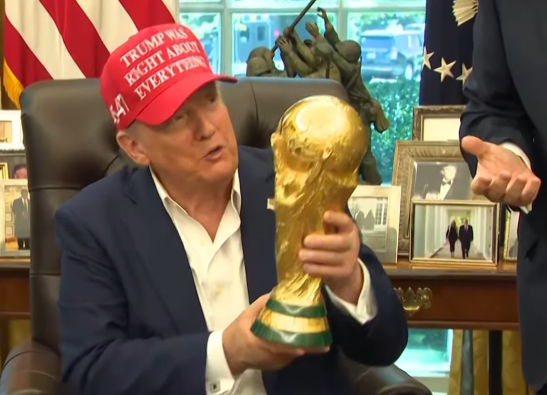 Donald Trump sits behind the Resolute Desk in the Oval Office holding a World Cup trophy replica while wearing a red baseball cap. Framed photographs, US flags and office furnishings can be seen in the background.