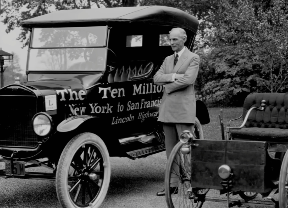 Henry Ford standing beside an early Ford automobile and a prototype vehicle, with “The Ten Millionth” car displayed behind him.