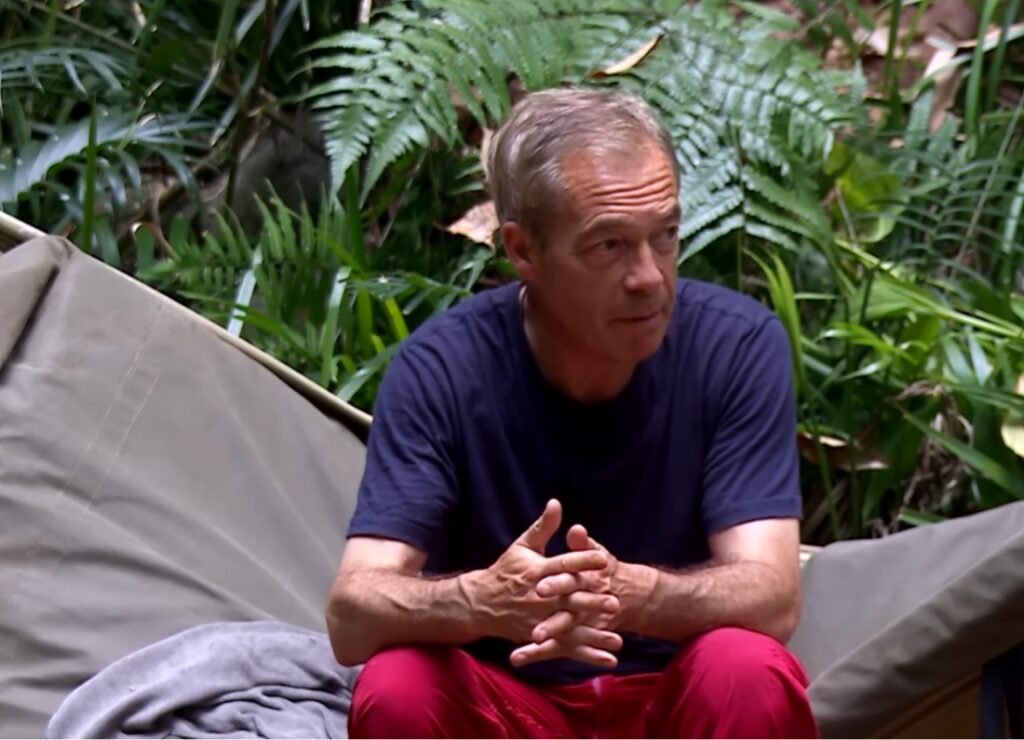 Nigel Farage sitting in the I’m A Celebrity… Get Me Out Of Here! jungle camp wearing a dark blue T-shirt and red trousers.
