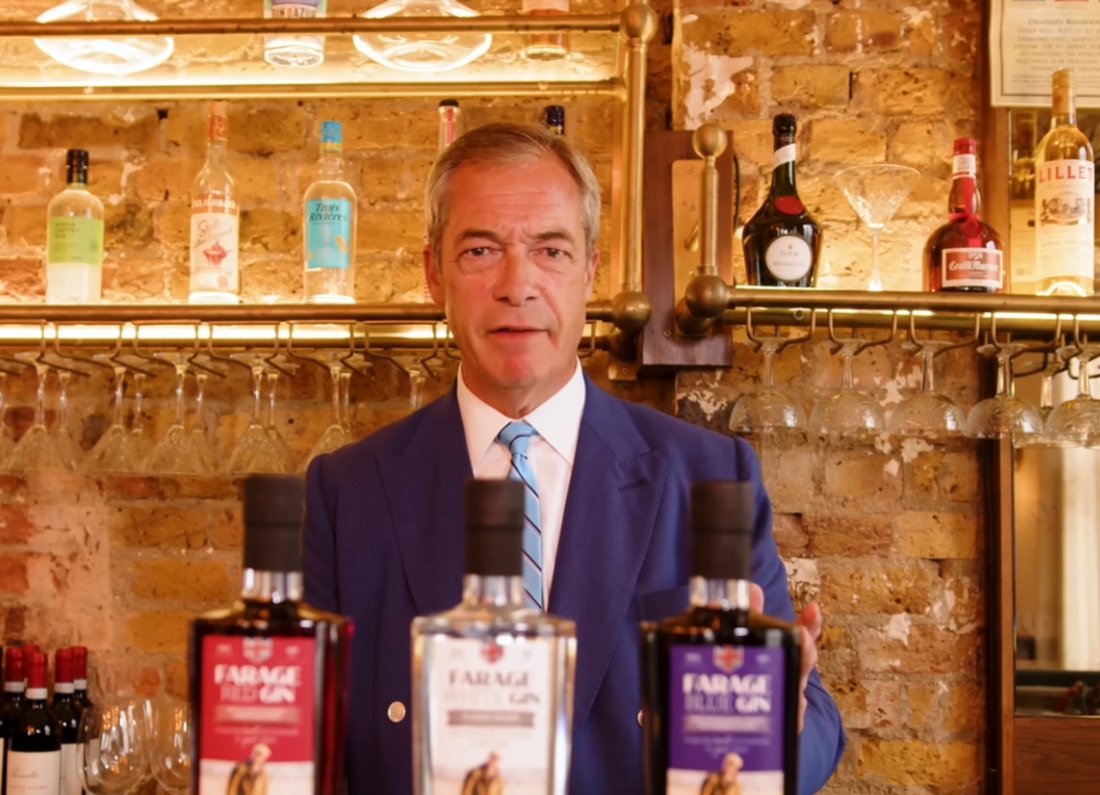 Nigel Farage stands behind a bar counter with bottles of Farage-branded gin displayed in the foreground. Shelves of spirits, wine glasses and exposed brickwork can be seen behind him.