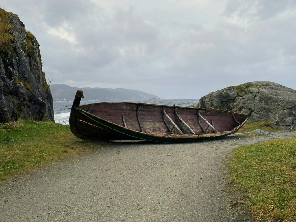 A wooden Viking-style boat rests on a coastal path between large rocks beside a rough sea under a cloudy sky.
