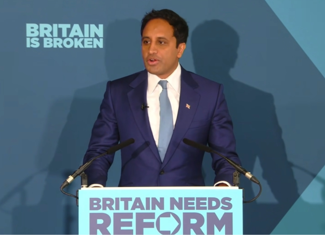 Zia Yusuf speaks at a Reform UK event with “Britain Needs Reform” displayed on the podium.