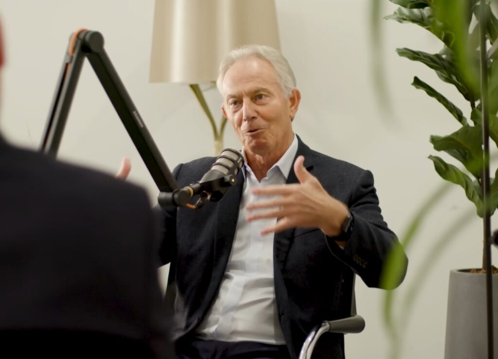 Former Prime Minister Tony Blair speaks into a podcast microphone while gesturing during an interview in a modern studio setting.