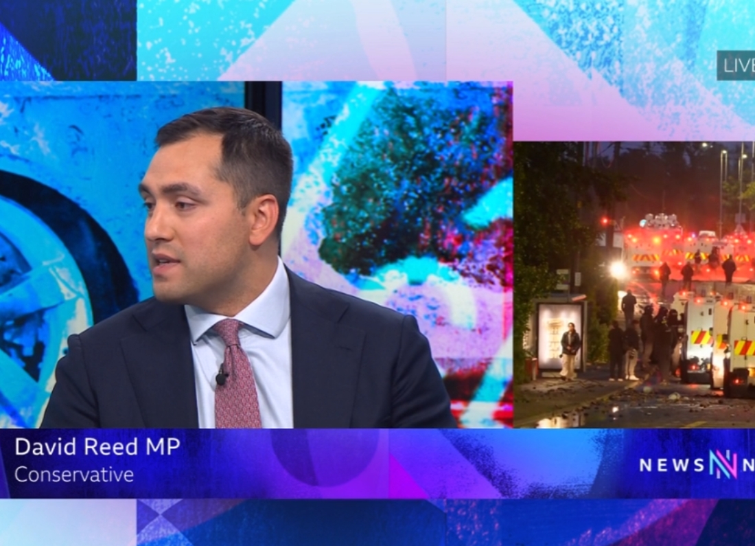 Conservative MP David Reed appears on BBC Newsnight during a discussion about the Belfast attack, with emergency service vehicles shown in an inset image beside him.