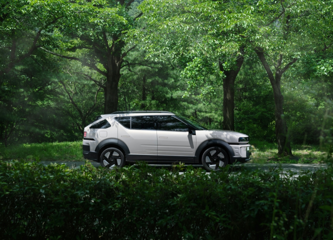 A modern electric SUV is parked on a road surrounded by dense green woodland, with sunlight filtering through the trees.