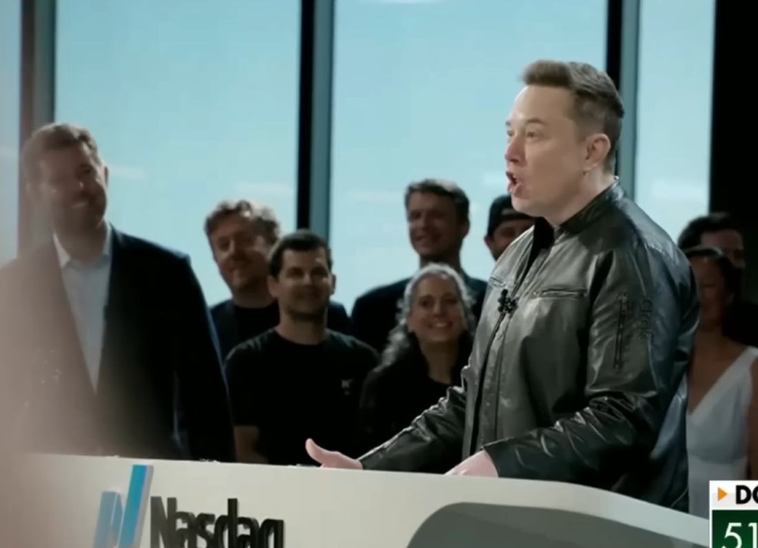 Elon Musk speaks from a stage at a SpaceX IPO event, wearing a black leather jacket and addressing an audience, with company employees and supporters visible in the background.