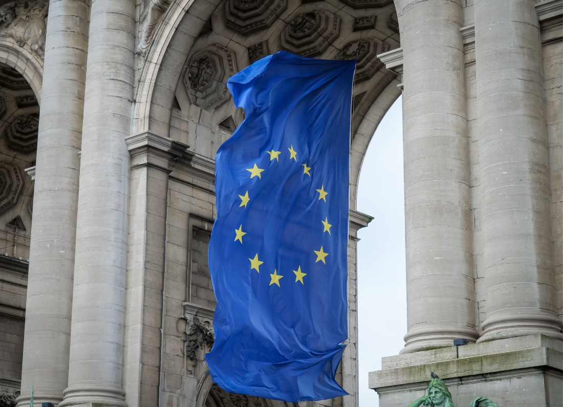 A large European Union flag hangs between towering stone columns of a grand historic building, billowing in the wind.