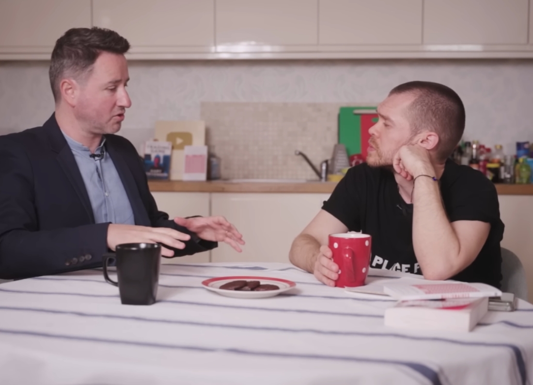 Two men sit at a kitchen table during a recorded discussion, with mugs, papers and a plate of biscuits in front of them.