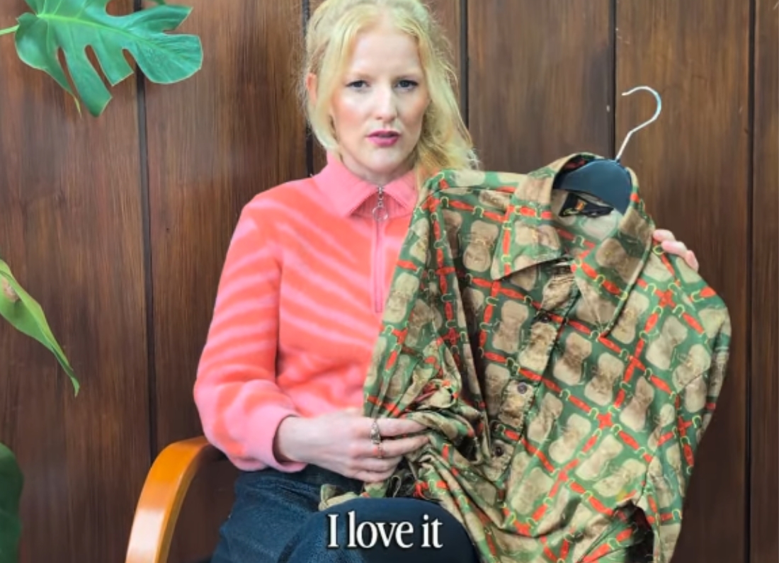Green Party MP Hannah Spencer sits indoors holding up a patterned shirt during a video interview, explaining that the garment was bought from a charity shop rather than a luxury fashion retailer.