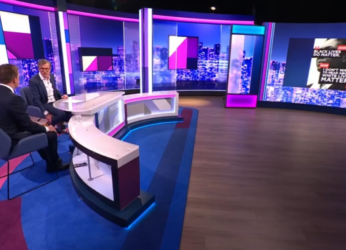 A television studio set during a political discussion programme, with presenters seated at a curved desk while graphics relating to political messaging and racism are displayed on a large screen.