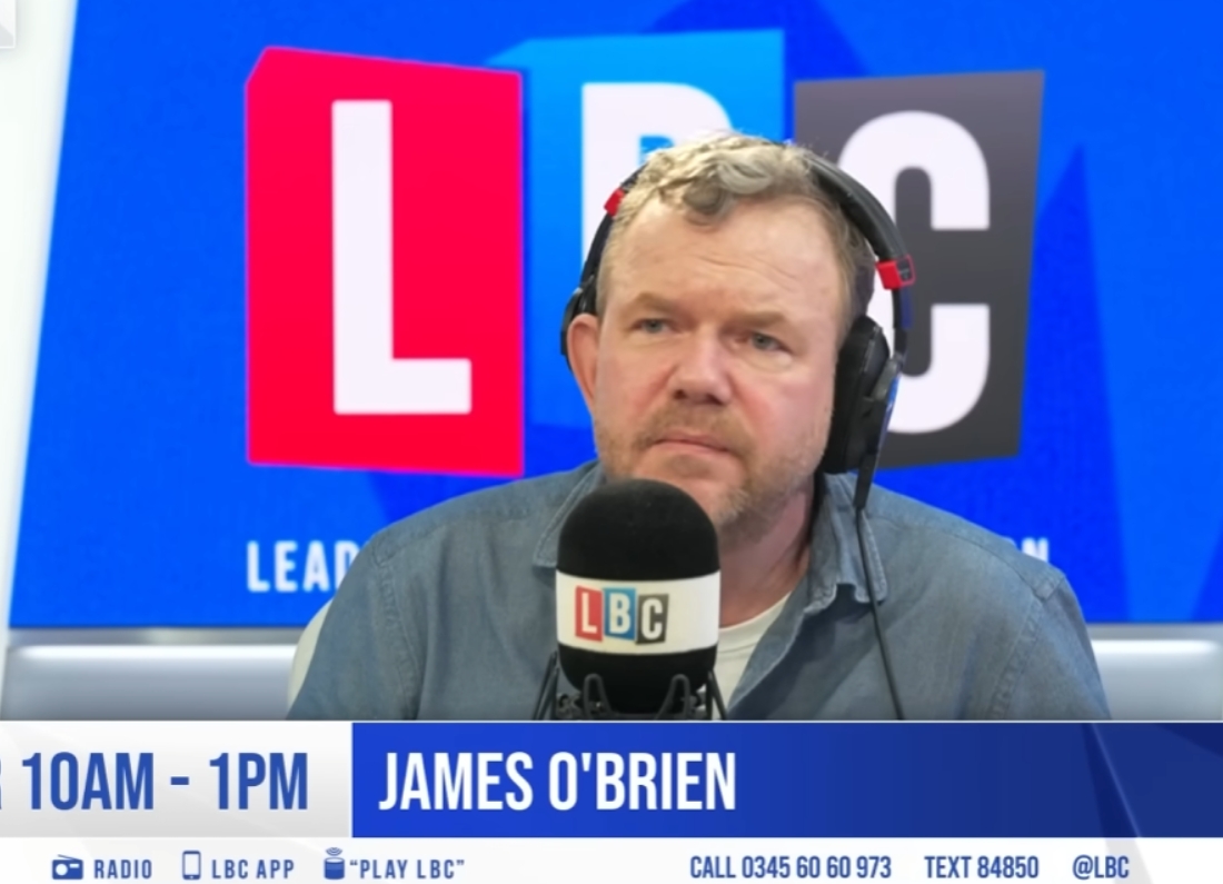 LBC presenter James O'Brien sits in a radio studio wearing headphones and speaking into a microphone during a live broadcast, with LBC branding visible in the background.