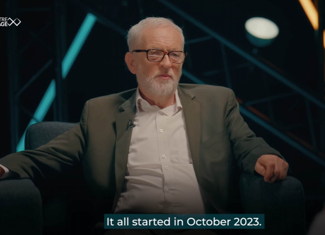 Jeremy Corbyn sits in an armchair during a filmed interview, speaking on a studio set with stage lighting in the background. A subtitle on screen reads: "It all started in October 2023."