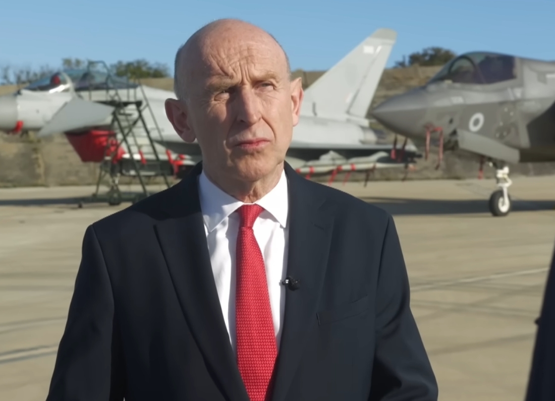 Defence Secretary John Healey speaks during an interview at a military airfield, with fighter jets visible in the background.