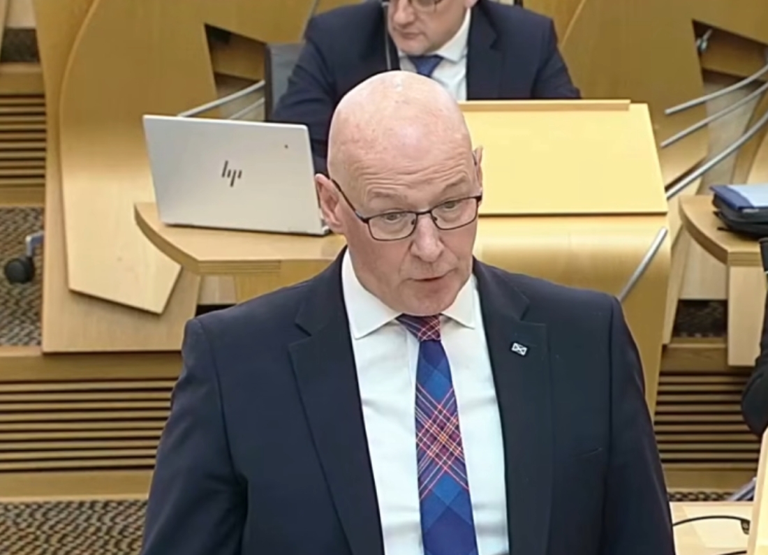 John Swinney speaks in the Scottish Parliament chamber while standing at his desk, wearing a dark suit and tartan-patterned tie.