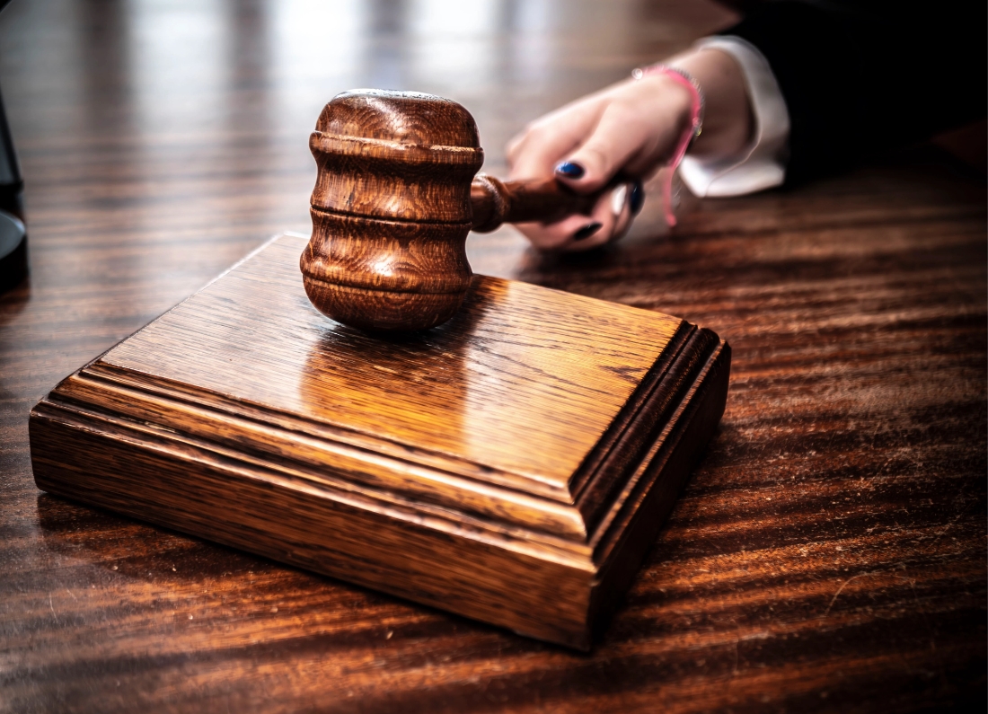 A close-up photograph of a wooden judge's gavel resting on a sound block, with a hand reaching toward it on a polished courtroom desk.