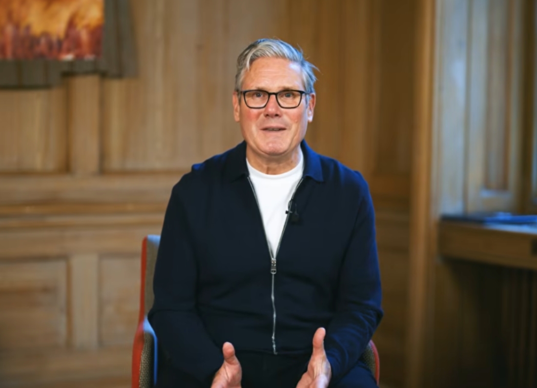 Prime Minister Keir Starmer sits indoors speaking directly to camera during a recorded address, wearing glasses and a dark zip-up jacket.