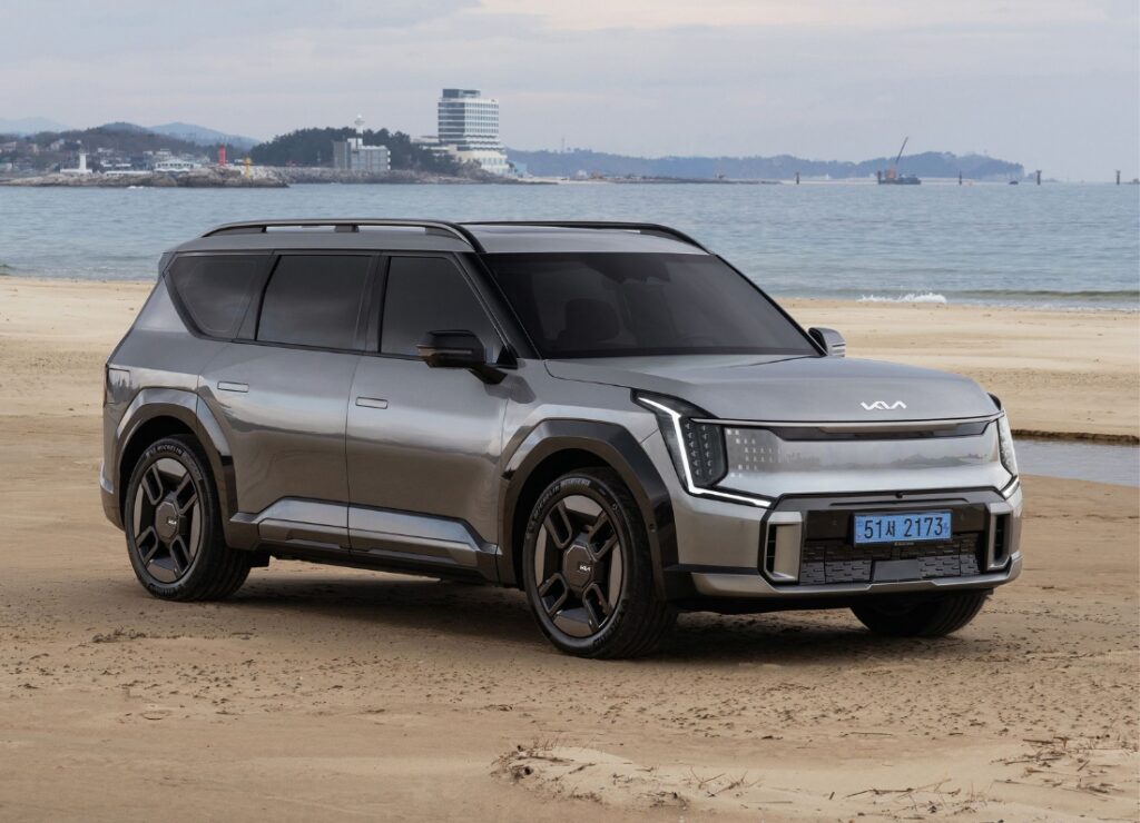 A Kia EV9 GT-Line electric SUV is parked on a sandy beach with calm water and a coastal skyline visible in the background.