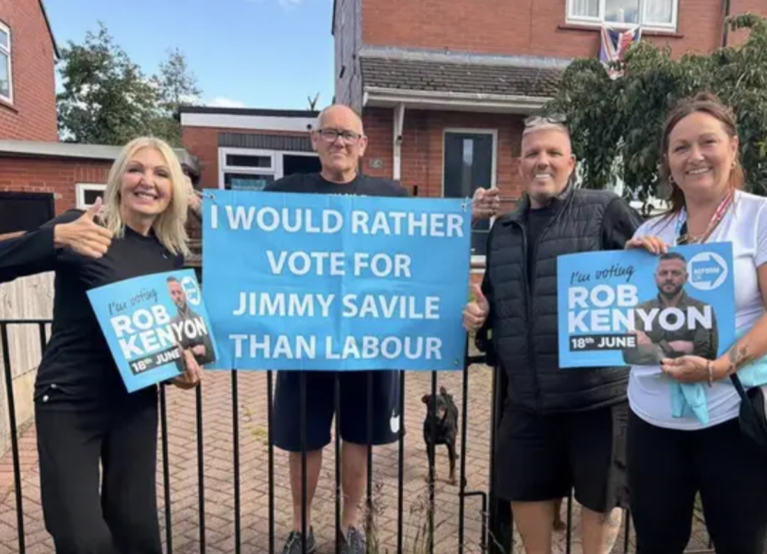 A group of Reform UK campaigners pose with election leaflets and a banner containing a controversial anti-Labour message during the Makerfield by-election campaign.