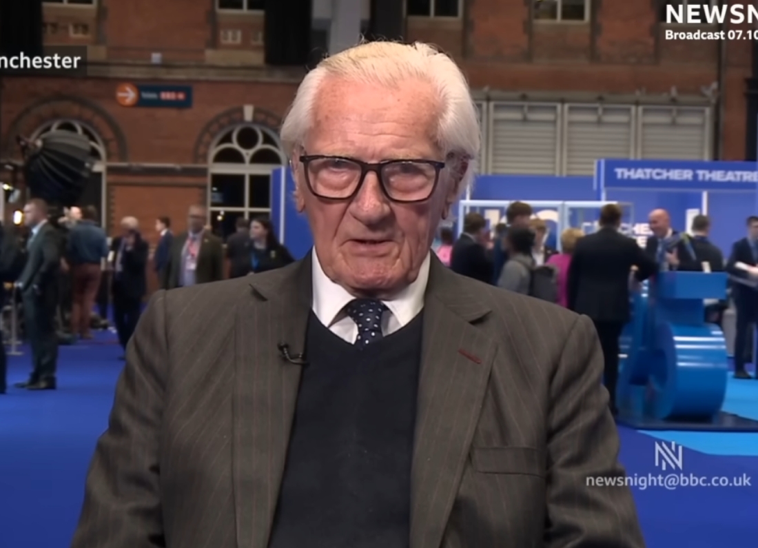 Lord Michael Heseltine appears live on BBC Newsnight from a Conservative Party conference venue in Manchester. The veteran Conservative politician is wearing a dark suit, navy jumper and tie, speaking directly to camera with delegates and exhibition stands visible behind him.