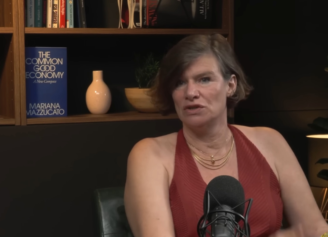 Economist Mariana Mazzucato speaking into a podcast microphone, with a copy of The Common Good Economy visible on a bookshelf behind her.