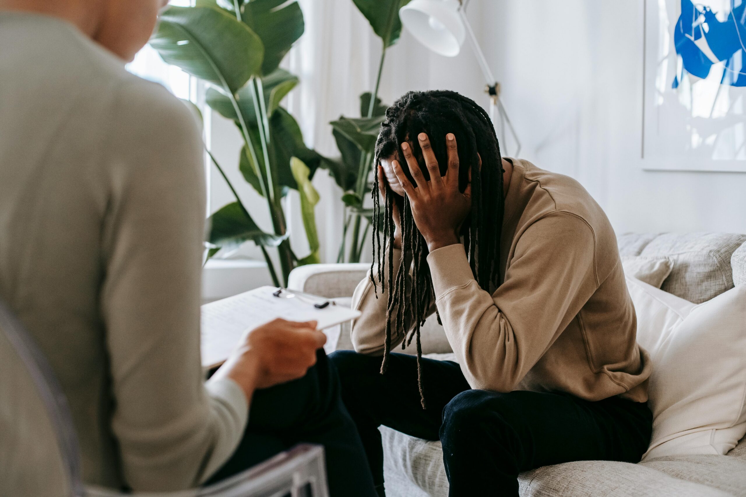A person sits on a sofa with their head in their hands during a counselling session, while another person holding a clipboard listens nearby.