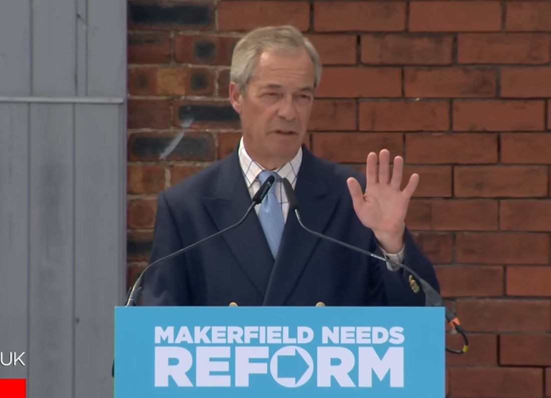 Nigel Farage speaks at a Reform UK campaign event from behind a podium carrying the slogan "Makerfield Needs Reform", raising one hand while addressing supporters.